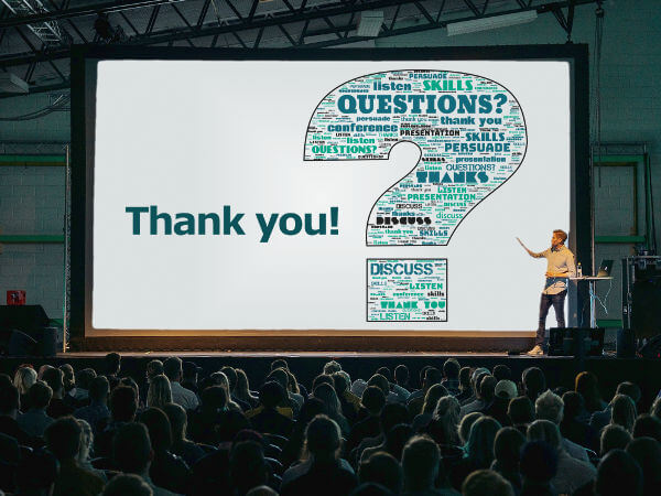 A presenter in front of a large audience, pointing at the screen displaying a presentation slide with a word cloud